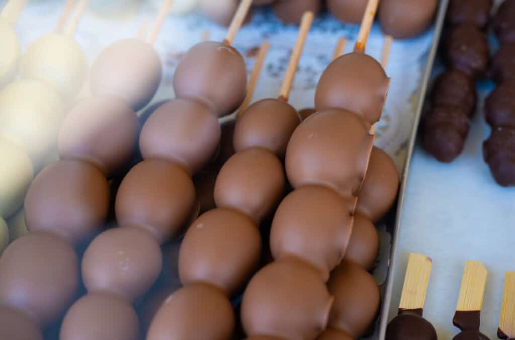 Mehrere Holzspieße mit mit Milchschokolade überzogenen Früchten, appetitlich präsentiert auf einem Marktstand.