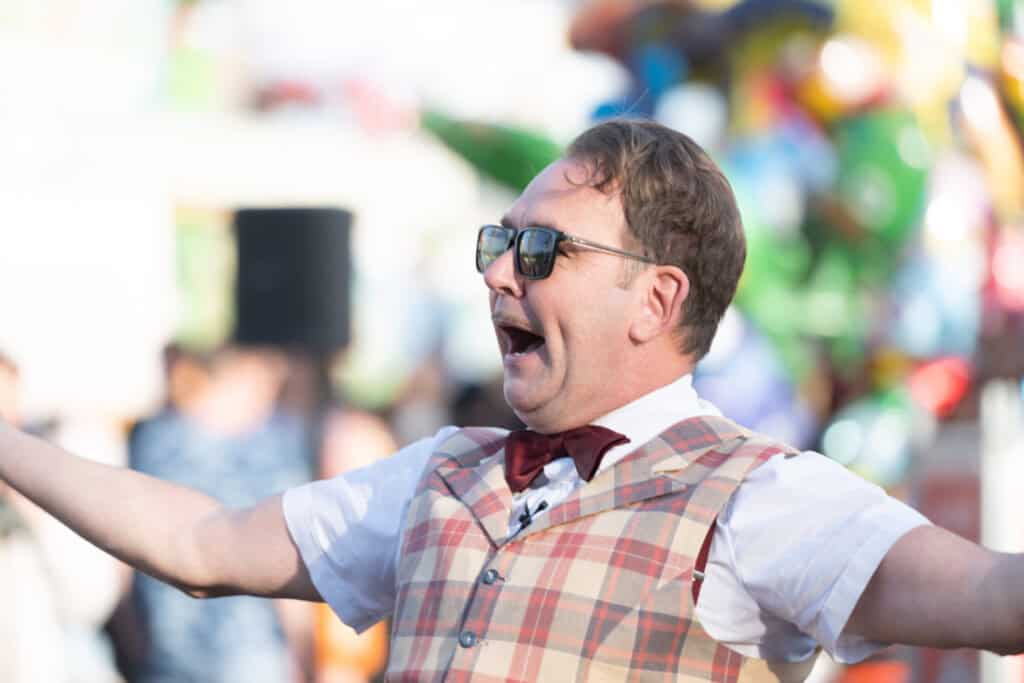 Schausteller in karierter Weste und roter Fliege in fröhlicher Pose bei einer Darbietung auf dem Volksfest.