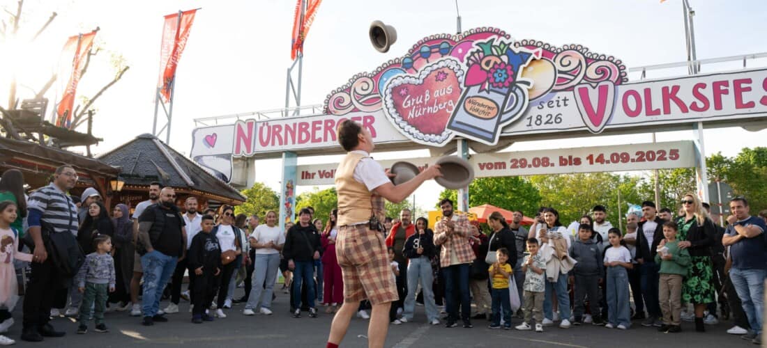 Jongleur in traditionellem Outfit unter dem dekorierten Eingangsportal des Nürnberger Volksfests mit Publikum im Hintergrund.